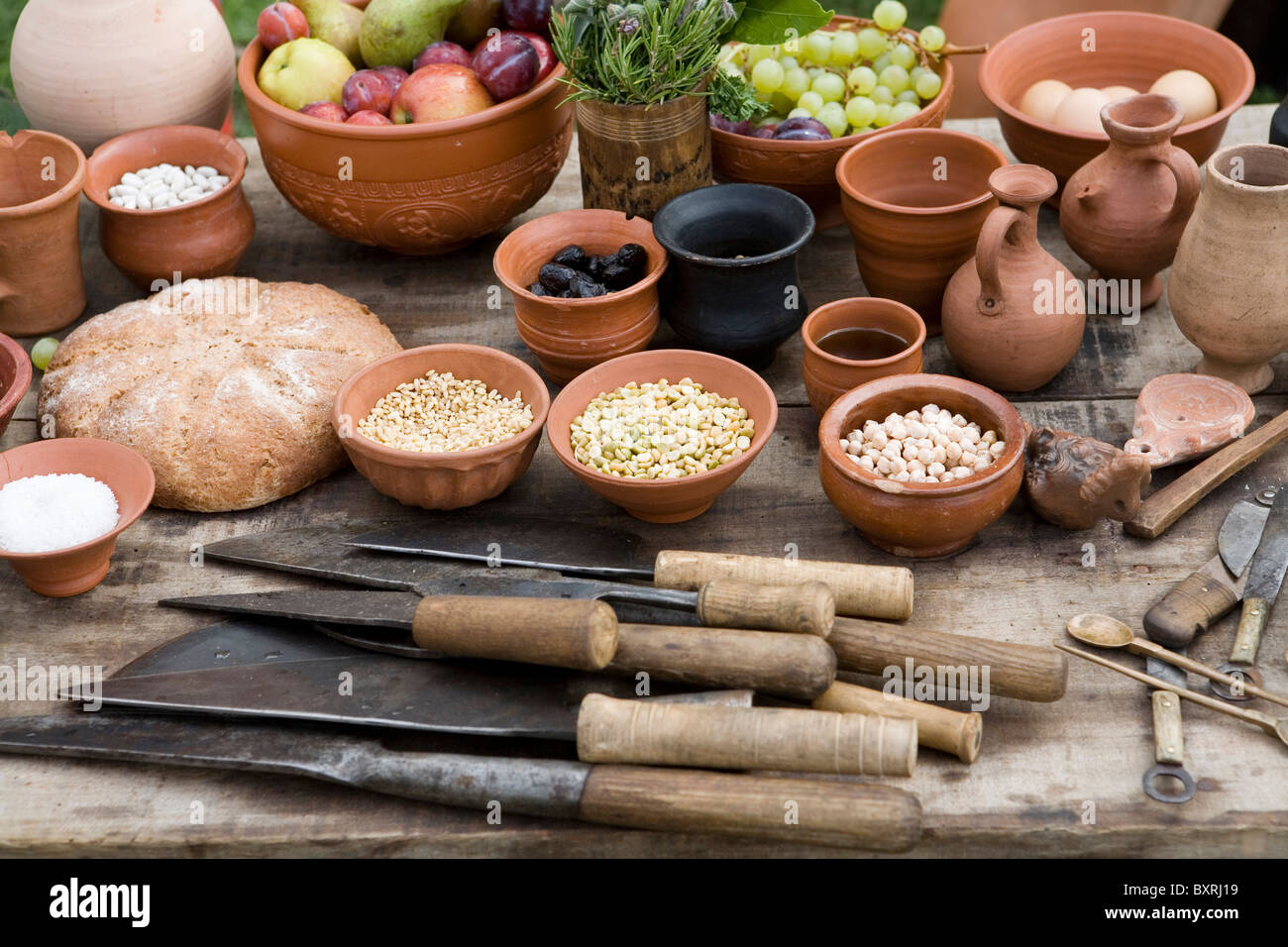Foods eaten by Roman soldiers, fruit, eggs, grains, and bread in terracotta pots and jugs with knives and spoons on table Stock Photo