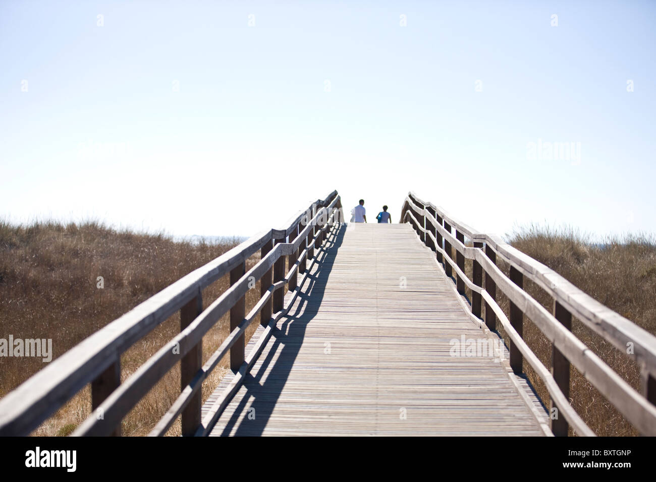 A wooden bridge over sand dunes, two people in the distance Stock Photo