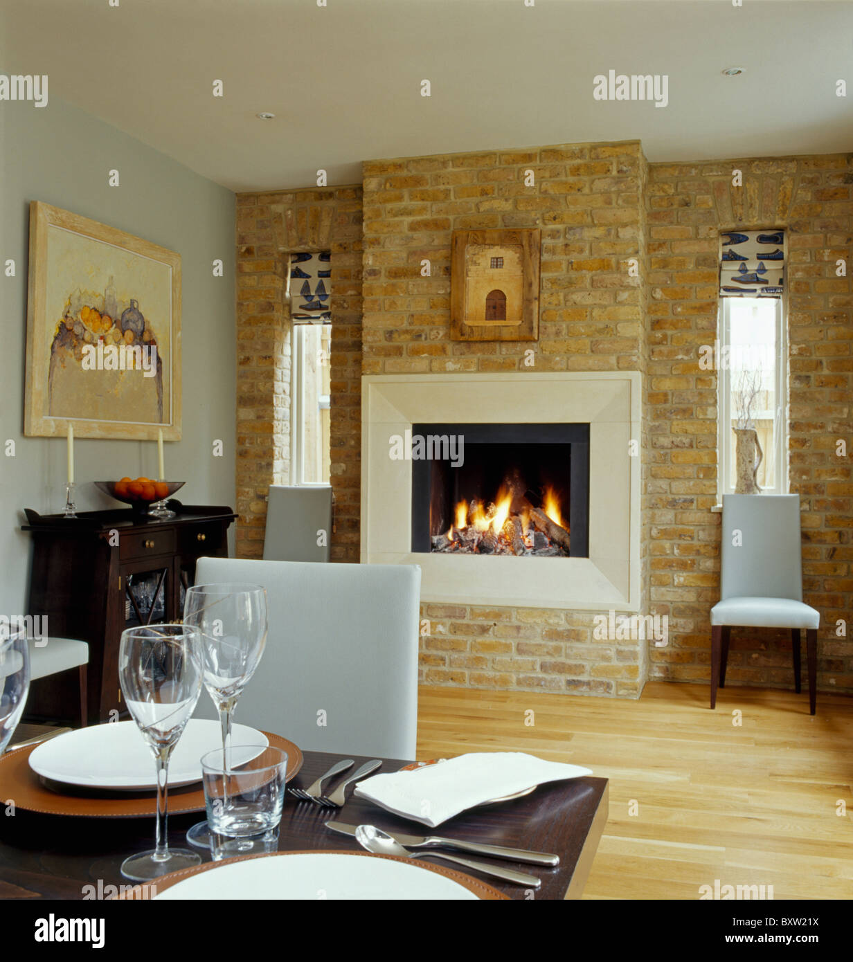 Lighted fire in fireplace in exposed brick wall in modern dining room with white leather dining chairs and wooden flooring Stock Photo