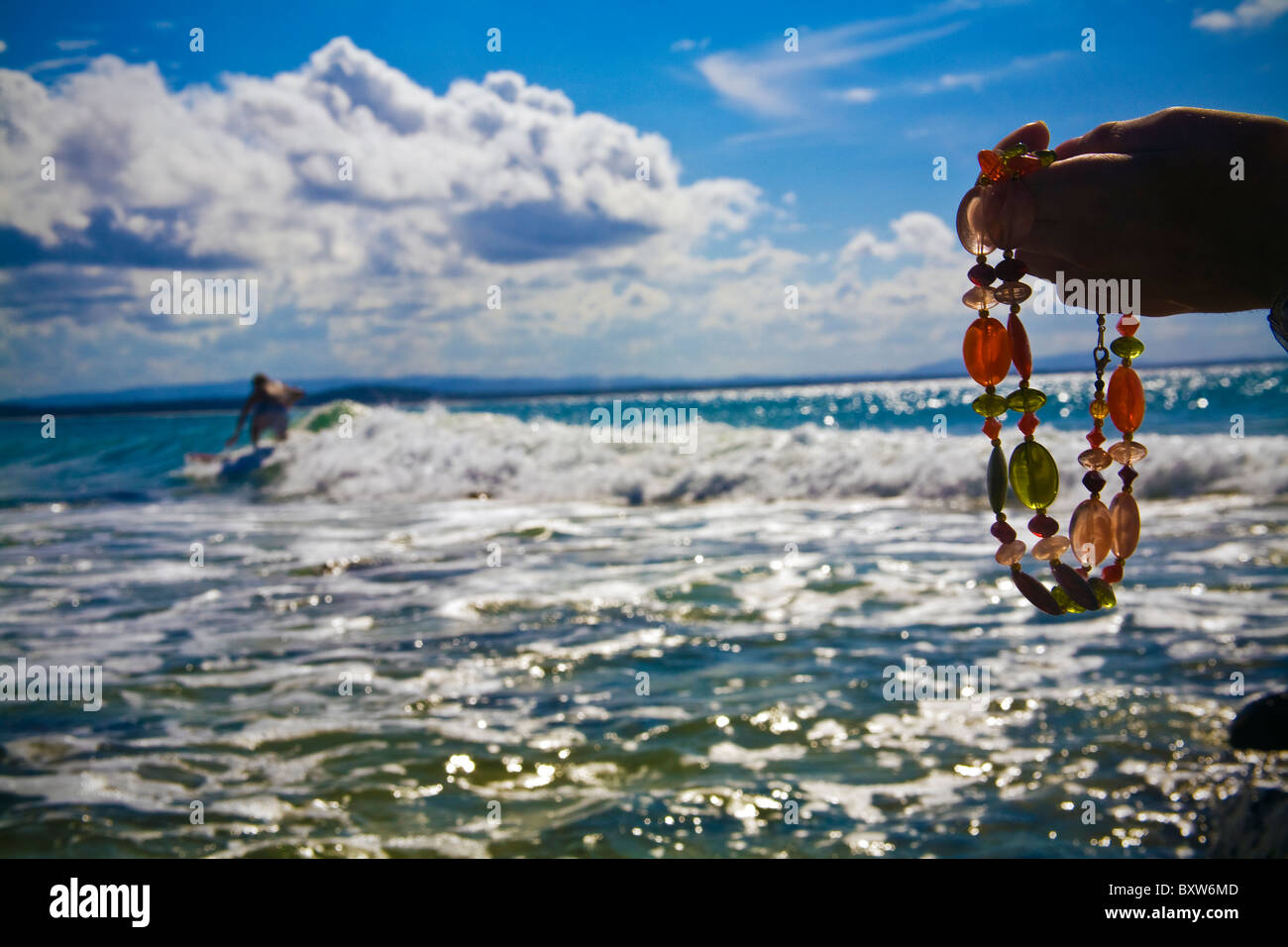 A woman's hand holding beaded necklace with Australian surfer riding a wave in background, beautiful blue sky day with clouds Stock Photo