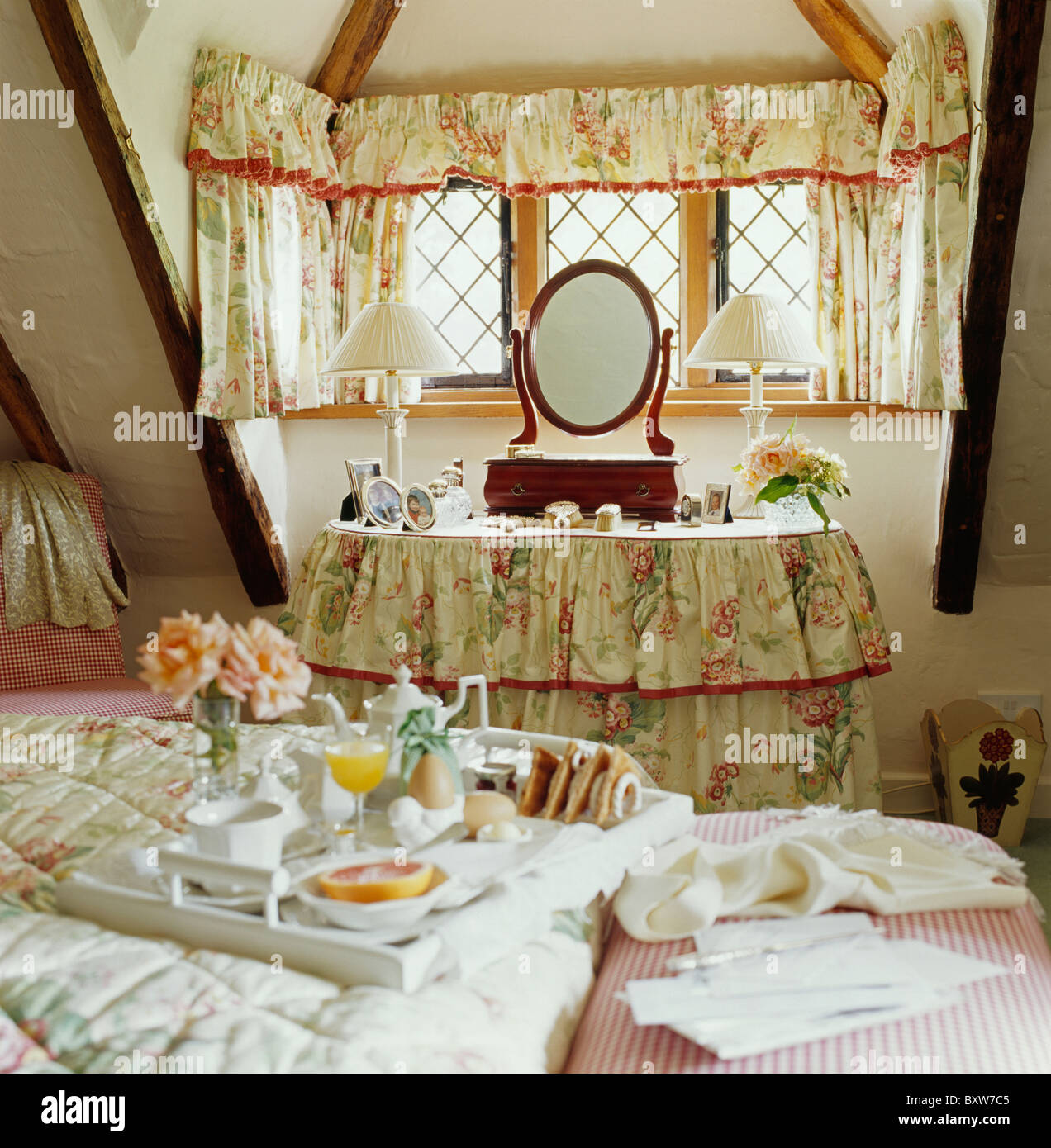 Floral curtains on window above dressing-table with matching drapes in cottage bedroom with breakfast tray on bed Stock Photo