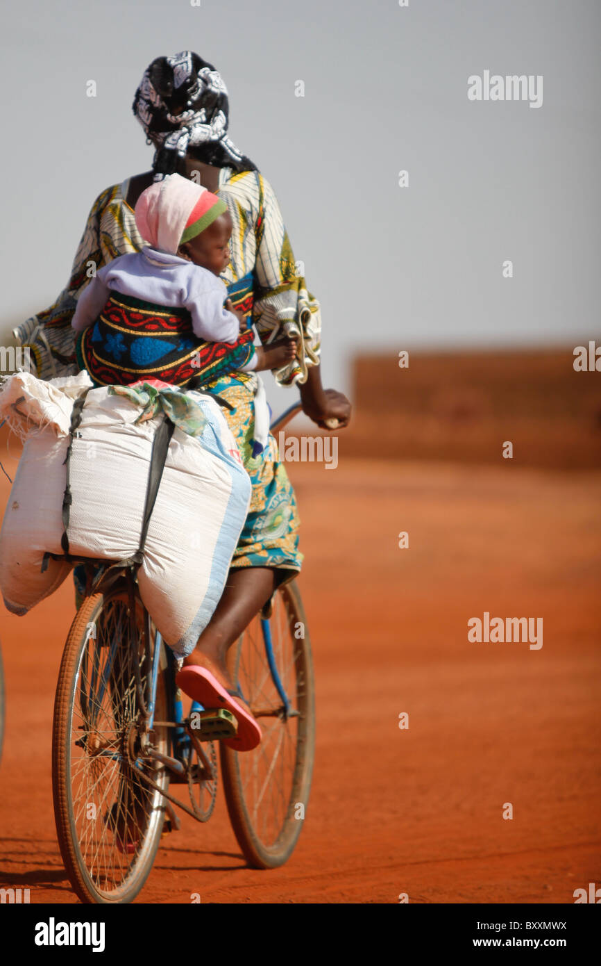 In Djibo in northern Burkina Faso, a woman carries her young child strapped to her back as she rides through town on a bicycle. Stock Photo