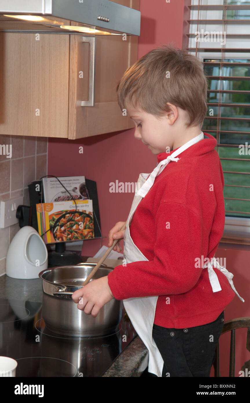 boy cooking making cakes standing on a chair next to the hotplates and oven wearing an apron. Child, Four-year-old Stock Photo