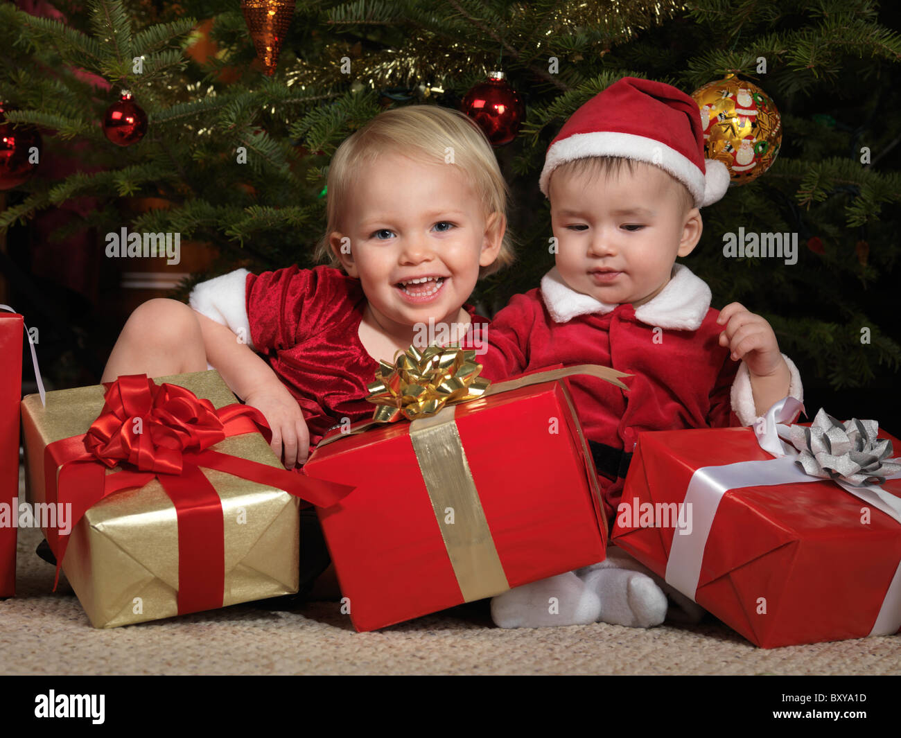 Two year old girl and an eight month old boy sitting with presents under a Christmas tree Stock Photo