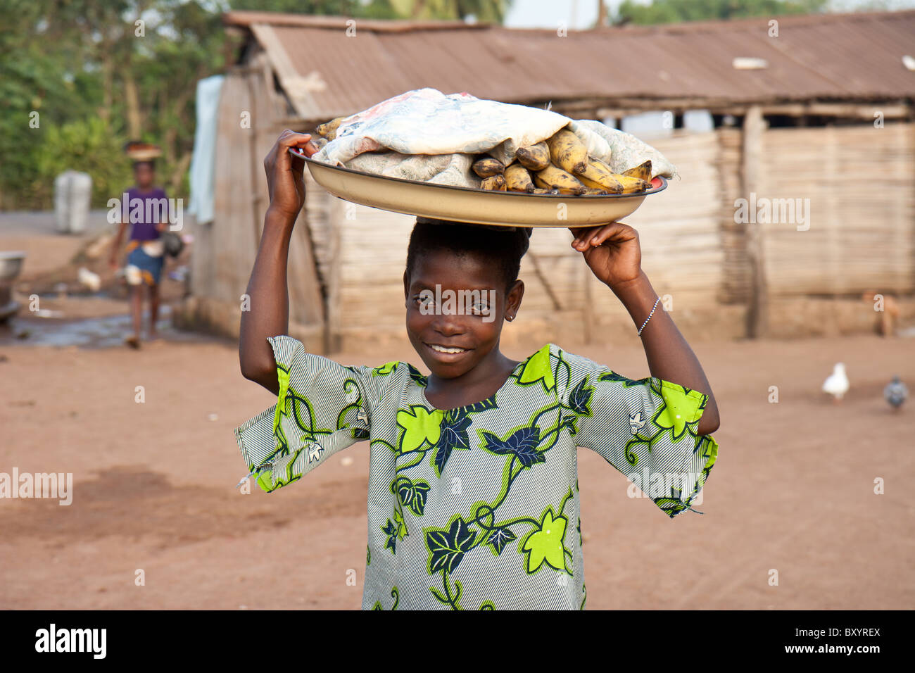 A young girl dressed in traditional pagne walks to a local market to sell plantain with her produce on her head. Stock Photo