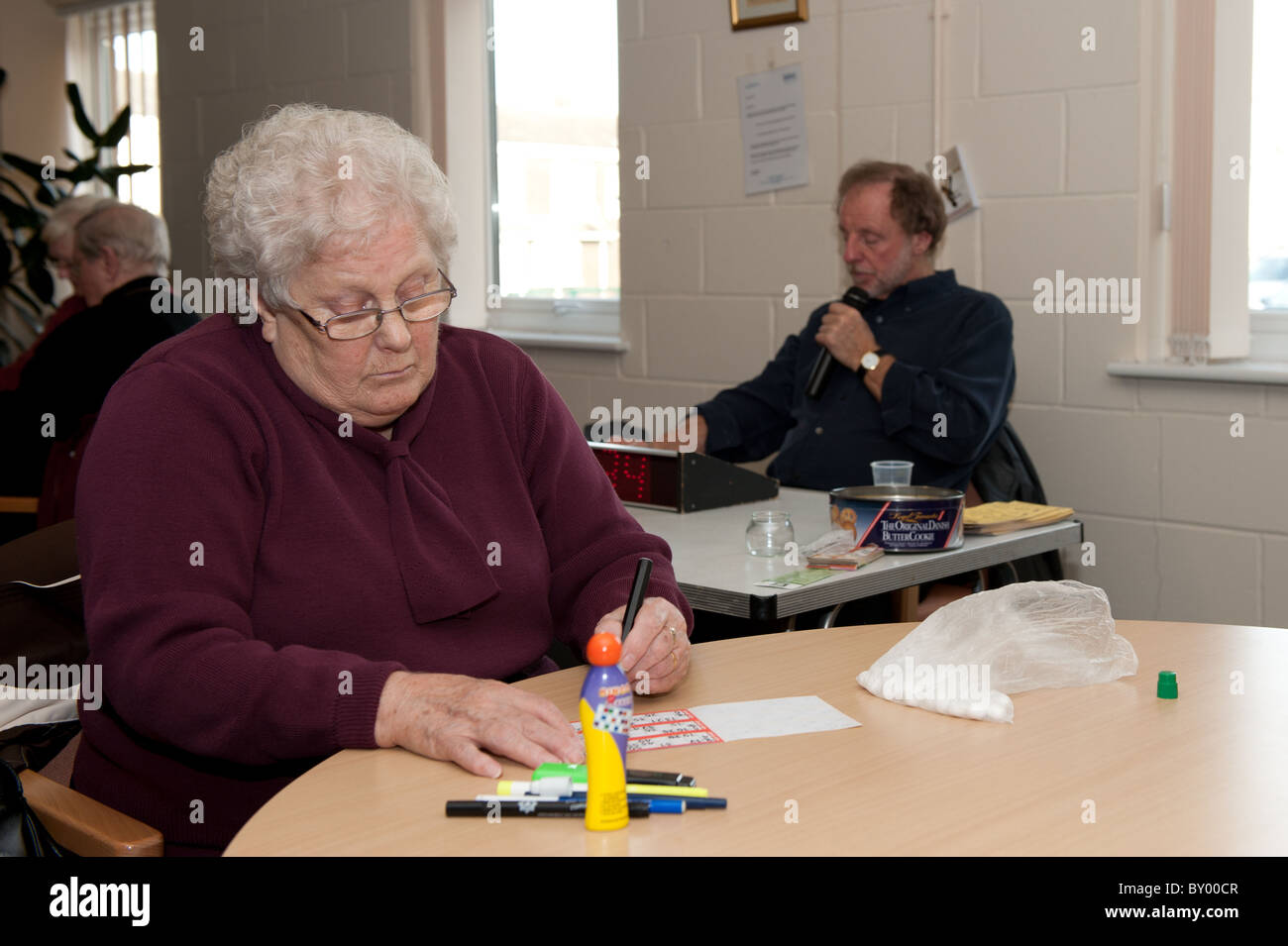 Elderly lady sat at table playing bingo in a day centre for the over 50s with volunteer bingo caller in the background. Stock Photo
