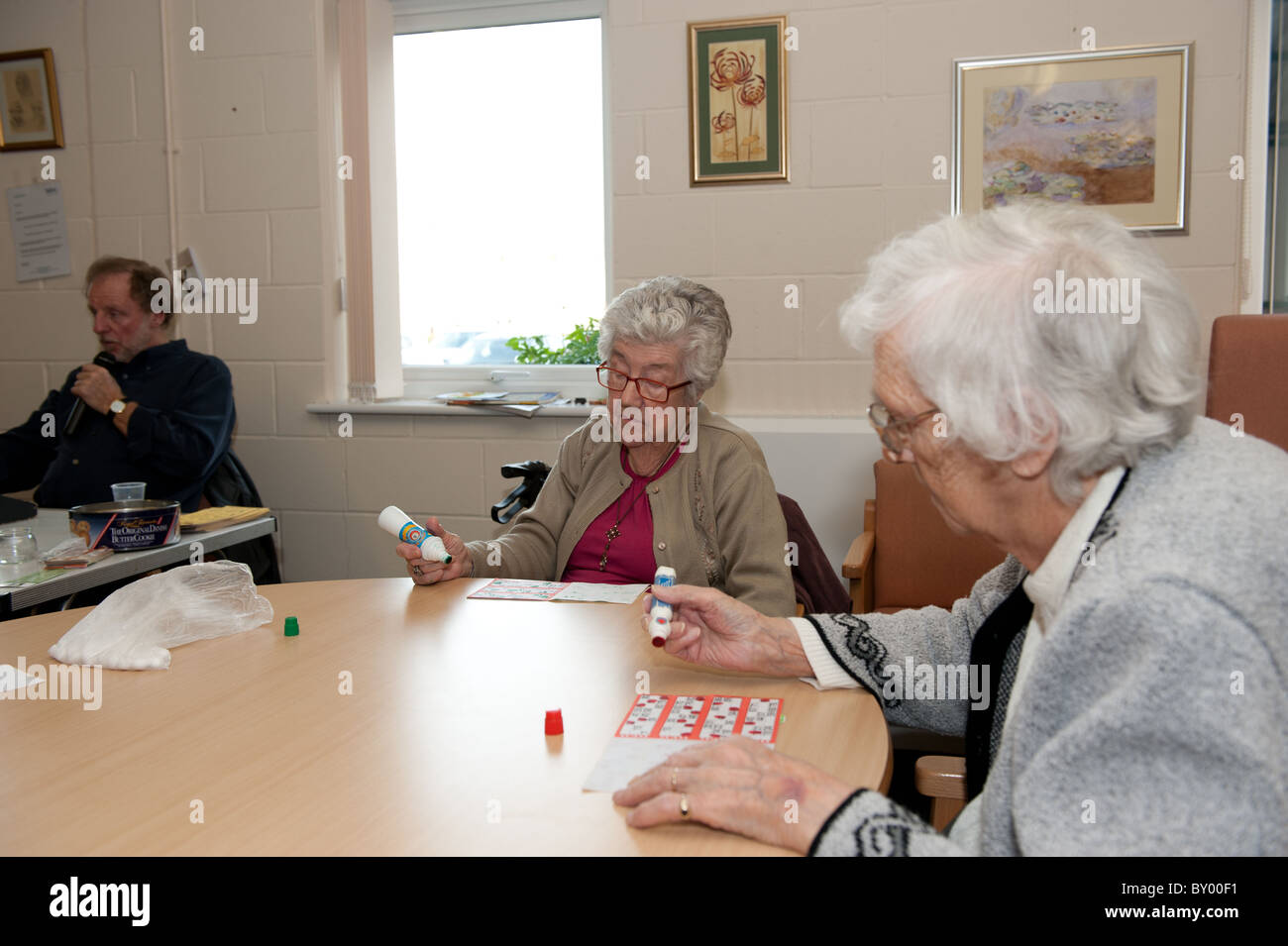 Two elderly ladies sitting at table in council run day centre playing bingo. Bingo caller visible in the background. Stock Photo
