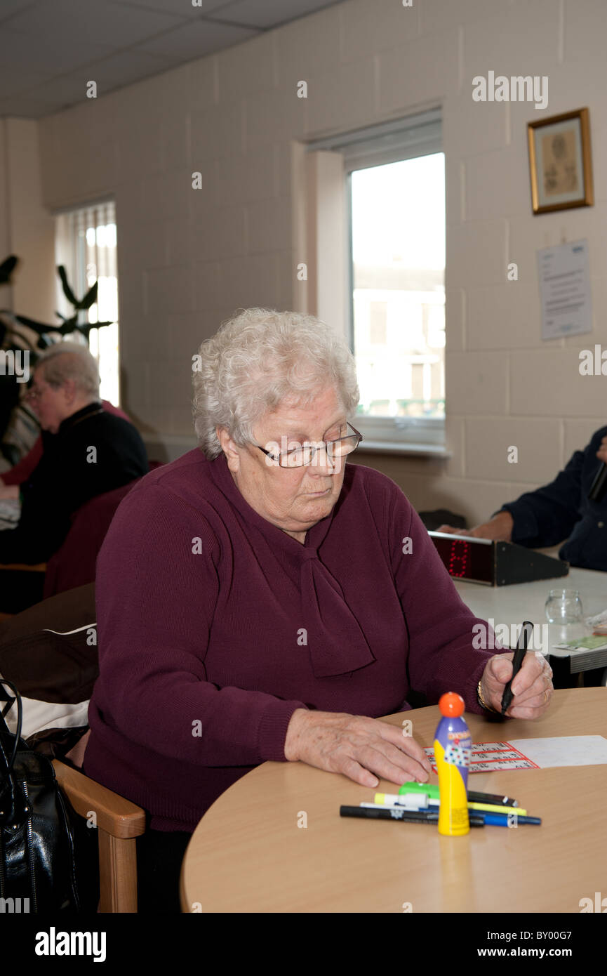 Elderly lady sat at table playing bingo in a day centre for the over 50s with volunteer bingo caller in the background. Stock Photo