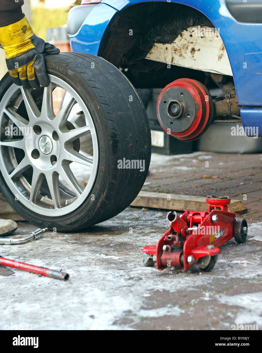 A mechanic removing a wheel from a car, held up by a jack on an icy driveway. Stock Photo