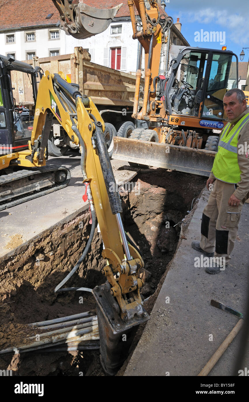 Workman supervising digging equipment excavating hole in road to repair power cables Saulieu France Stock Photo