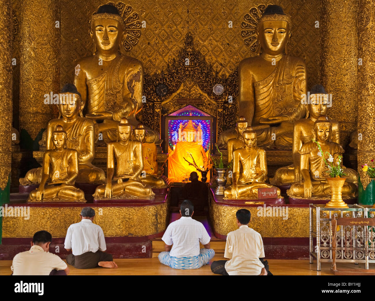 Myanmar, Burma, Yangon, Rangoon. Devout worshippers praying in front of golden Buddha statues, one with a halo of radiating Stock Photo