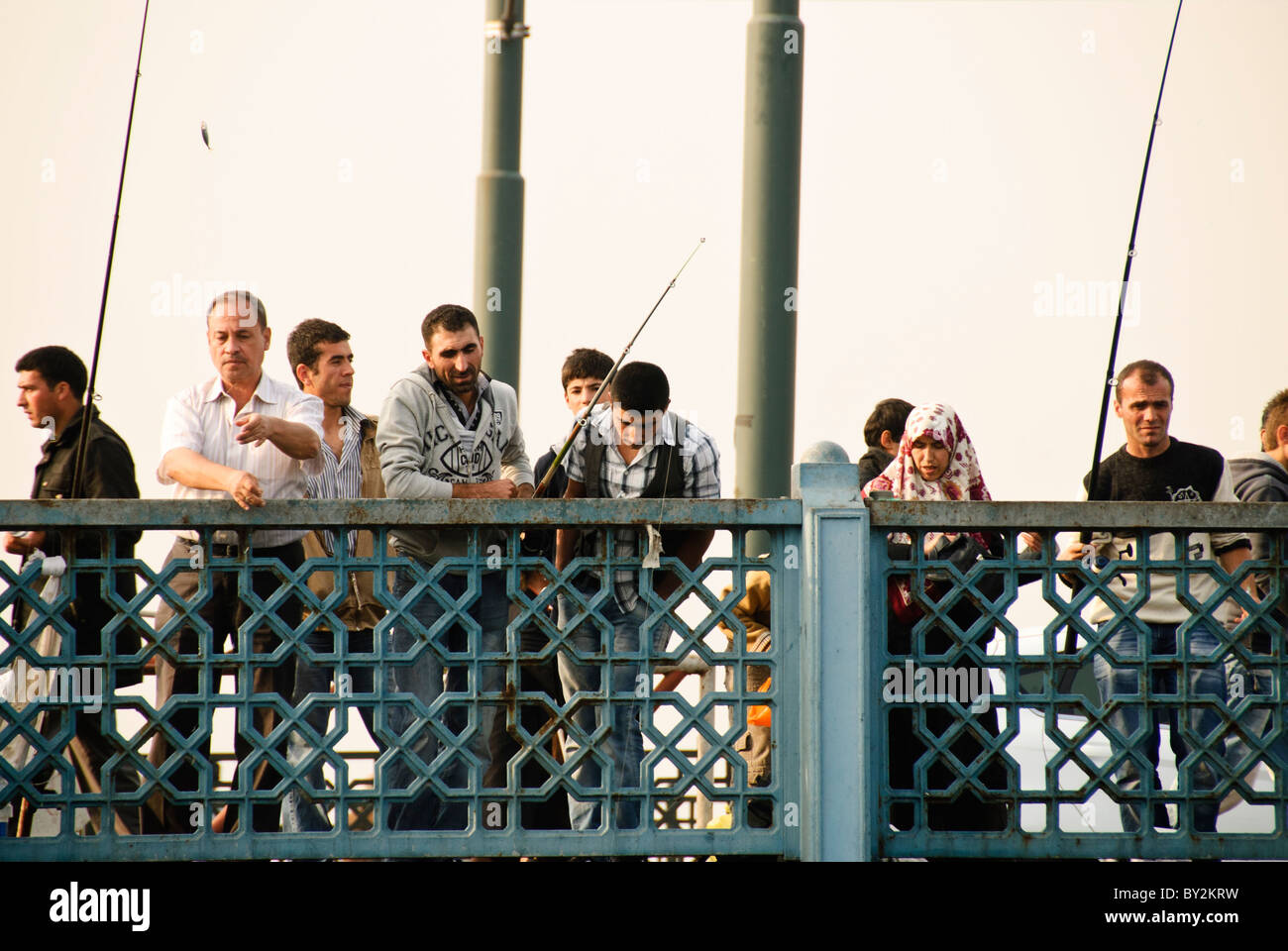 Galata Bridge Fishing Istanbul Turkey // ISTANBUL, Turkey — Fishing off Istanbul's historic Galata Bridge spanning the Golden Horn. Several individuals, including men and women, are seen casting their lines, with one person actively reeling in a fish. Stock Photo