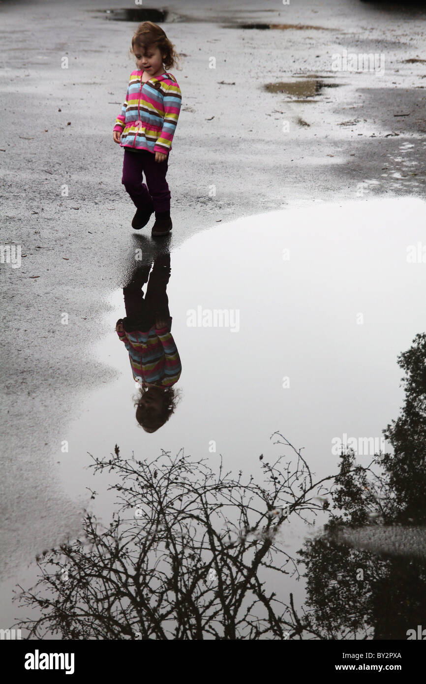 A 3 year old girl walking along the edge of a puddle, with tree branches reflected in it. Stock Photo