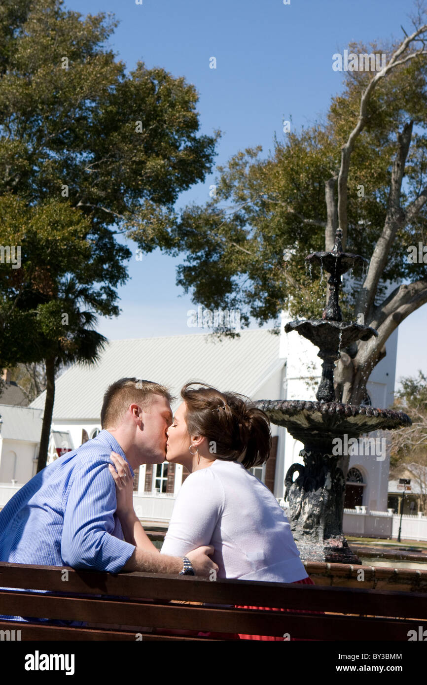 A young engaged couple sit on a park bench kissing by a water fountain. Stock Photo