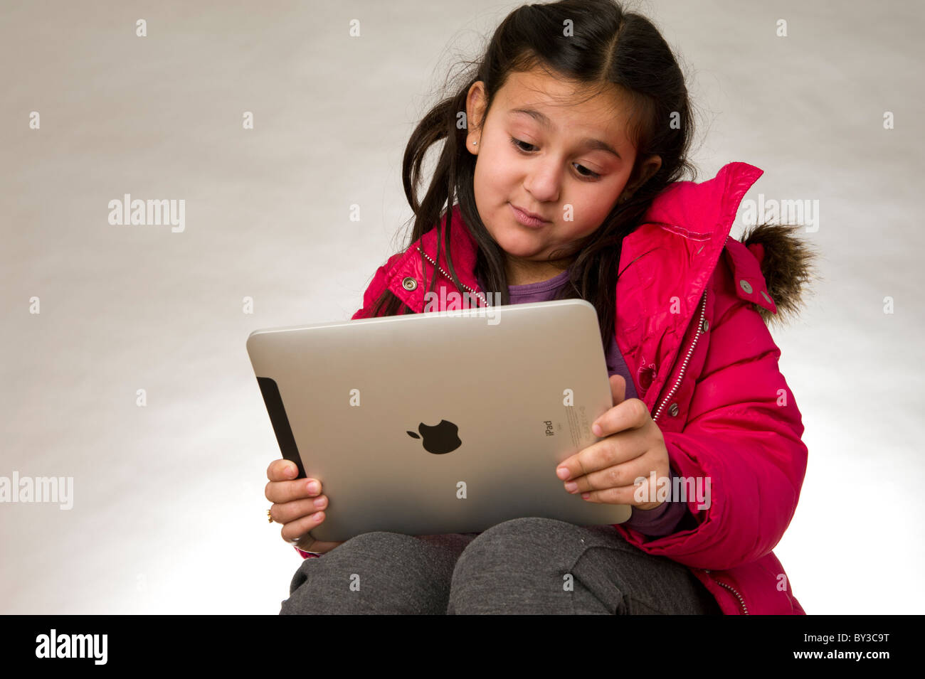 Studio shot of a young girl wearing a red coat and  playing a game on the touch pad screen of an ipad Stock Photo