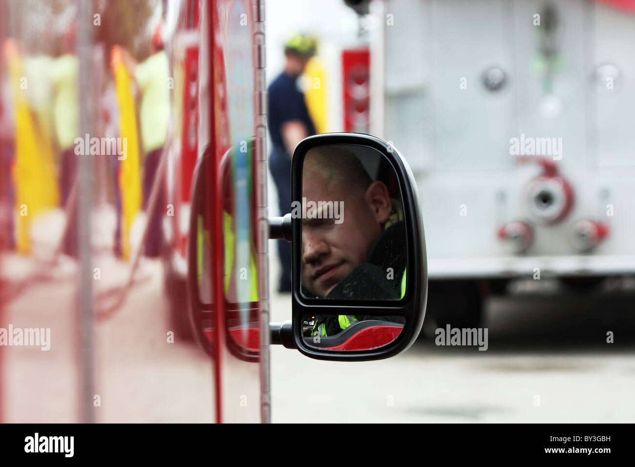 A firefighter reflection in the fire truck vehicle rear view side mirror and a firefighter in the background Stock Photo