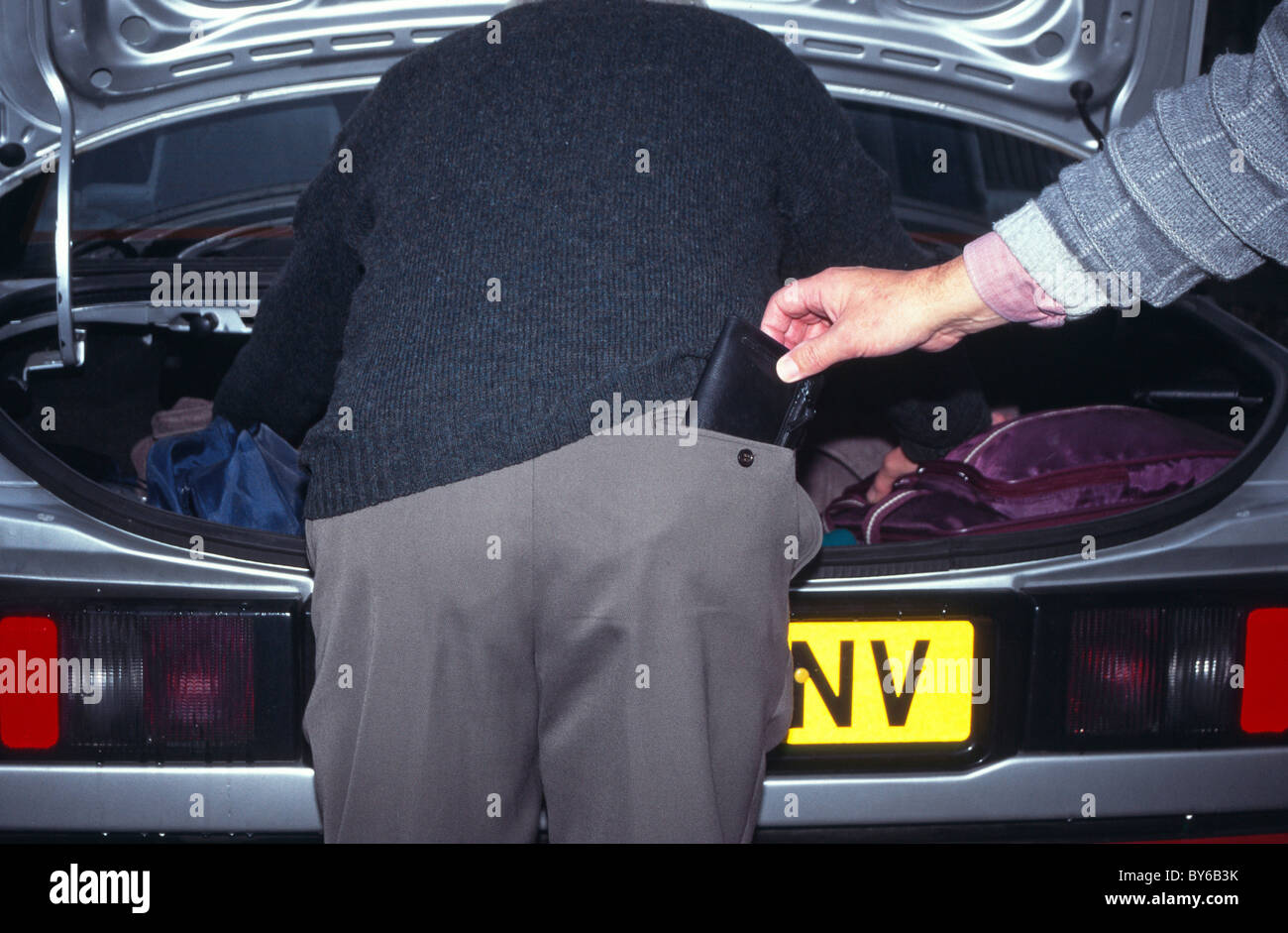 Pickpocket lifting wallet from old man back pocket in public car park whilst distracted looking in car boot posed by models England UK Stock Photo