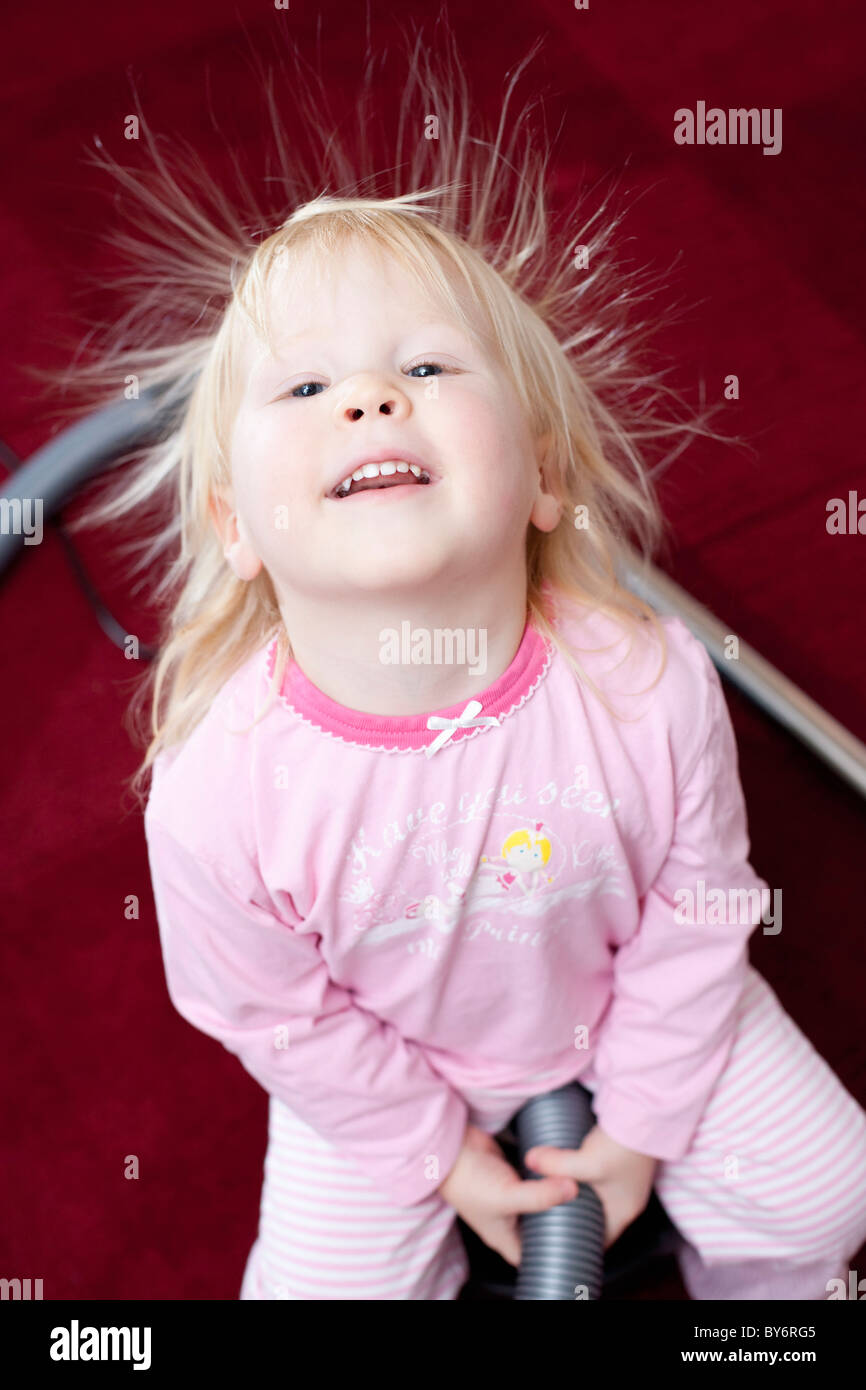 Little girl in pink clothes with spiky hair sitting on vacuum cleaner on red carpet, Germany Stock Photo
