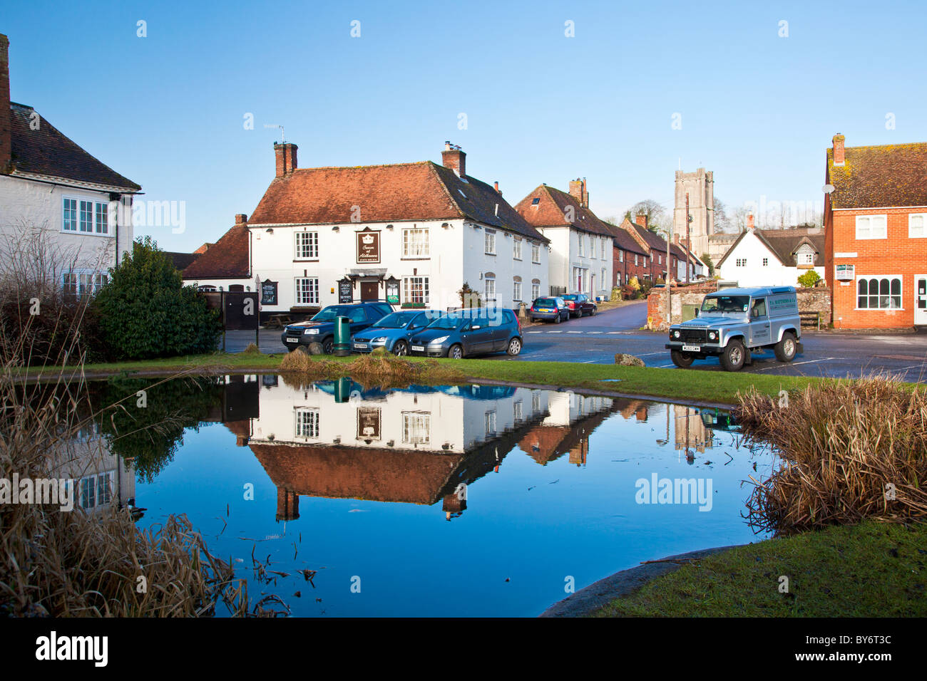The square, houses, pub and church reflected in the village pond in winter in the village of Aldbourne, Wiltshire, England, UK Stock Photo