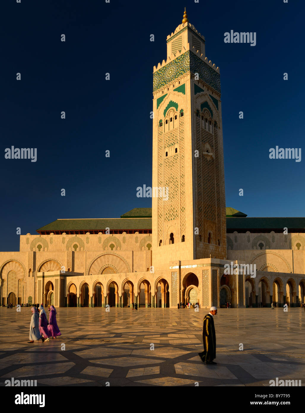Moroccan man and women walking on the plaza of the Hassan II Mosque Casablanca at sunset with minaret of Moorish architecture in Casablanca Morocco Stock Photo