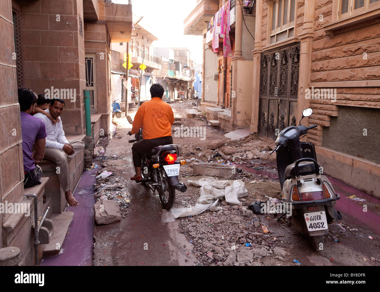 India, Rajasthan, Jodhpur, family sitting outside home in dilapidated street as motorcyclist rides past Stock Photo