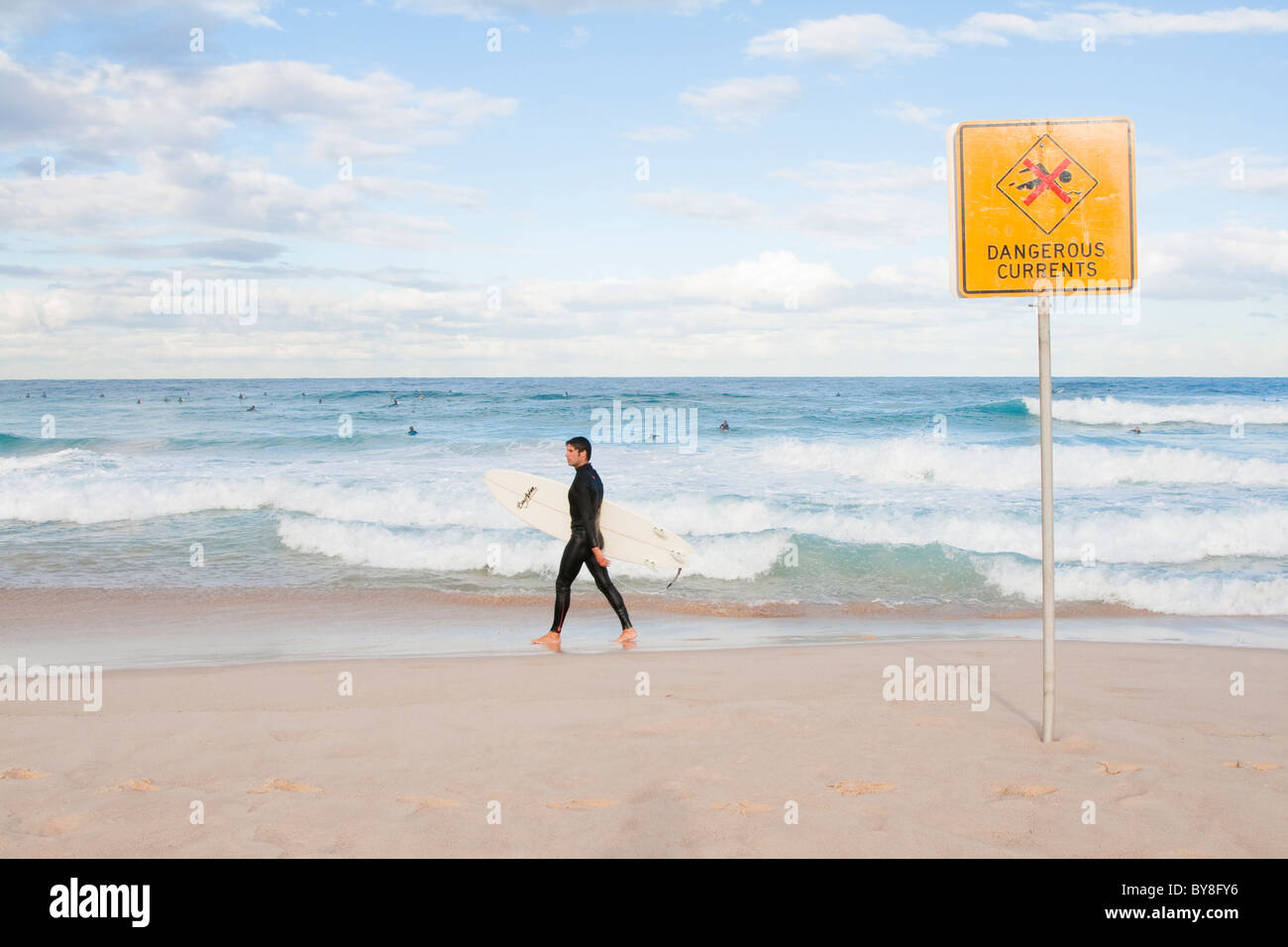 Male surfer walks along Bondi Beach, past signs warning of dangerous currents Stock Photo