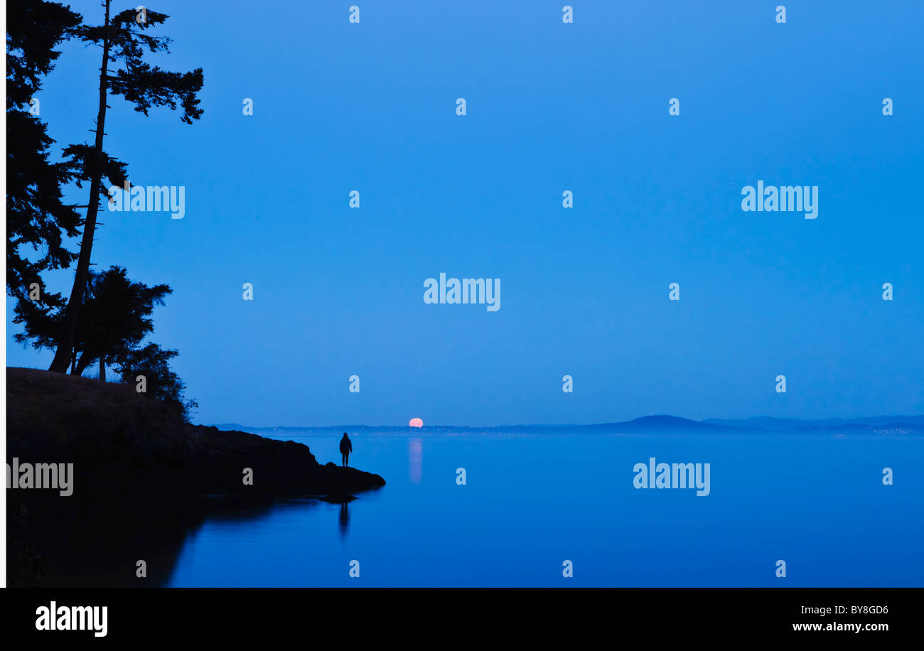A woman stands on a shoreline watching the setting full moon over Haro Strait and Vancouver Island, San Juan Island, WA, USA. Stock Photo