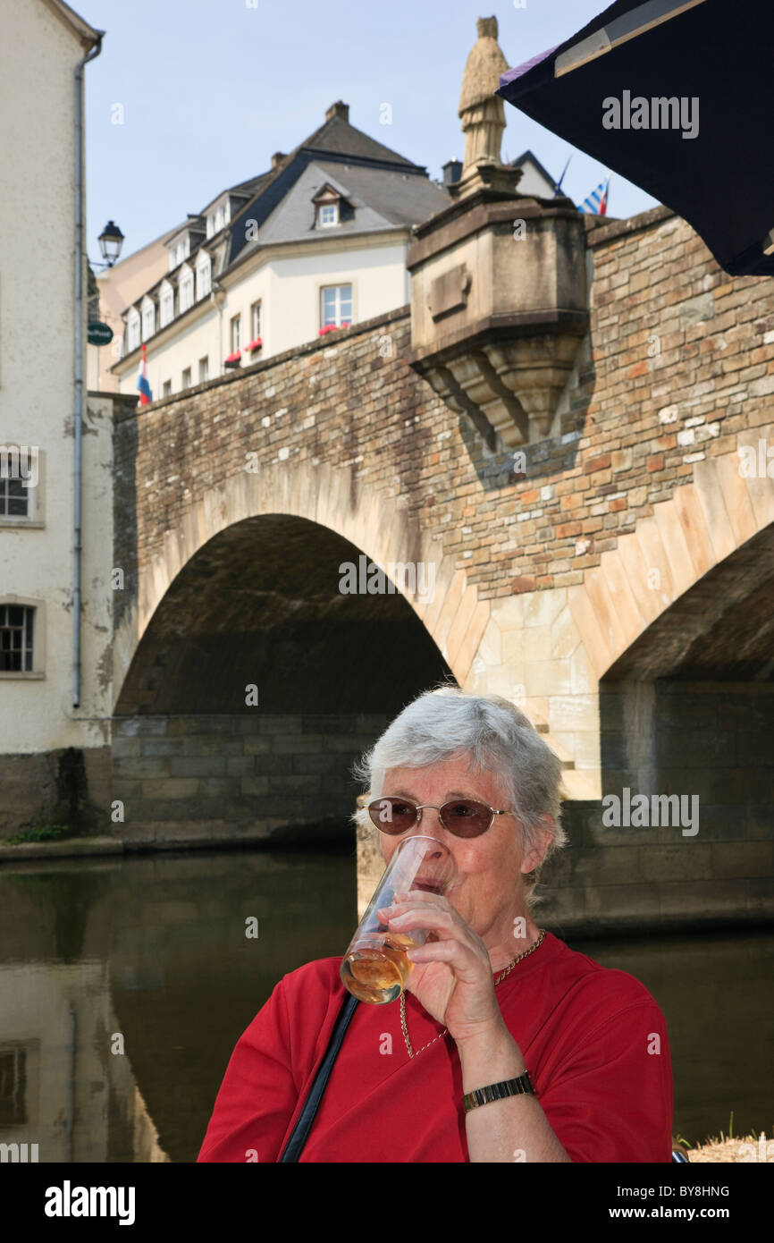 Senior woman tourist drinking a glass of beer in an outdoor cafe by bridge over River Our. Vianden, Grand Duchy of Luxembourg Stock Photo