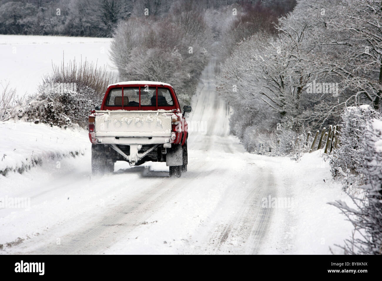 An old red Toyota pick up truck cautiously drives through the snow during a cold winter in England. 2010. Stock Photo