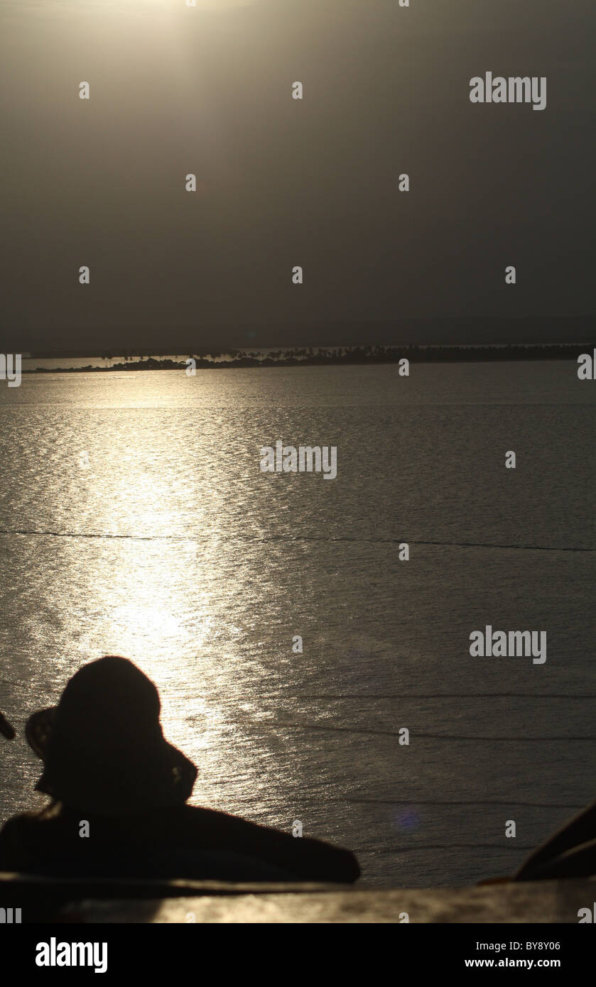 A woman sitting on a beach looks out at the sun setting over the sea ...