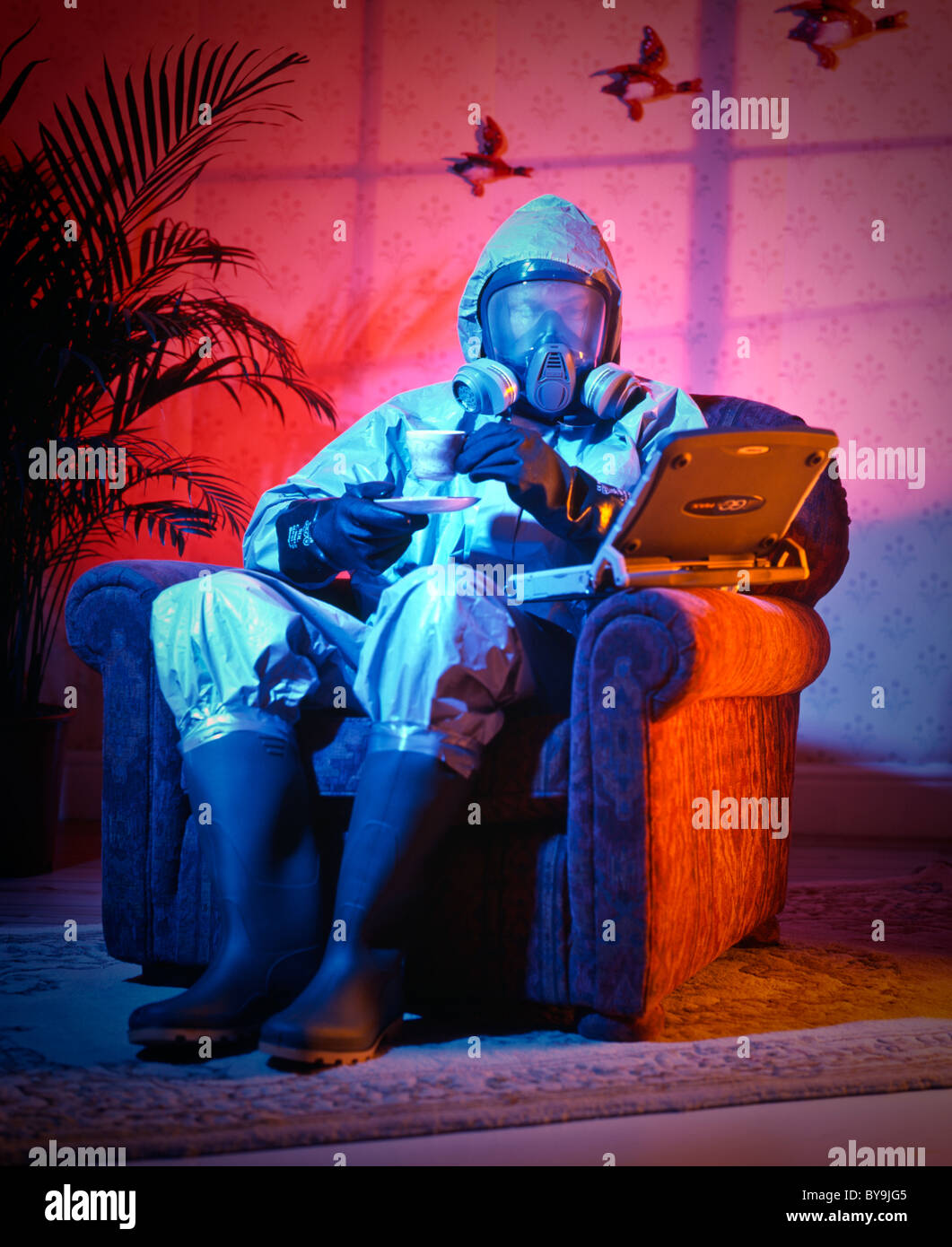 man in sitting in an arm chair in a suburban domestic location with his lap top and a cup of tea wearing a thermo-nuclear suit Stock Photo
