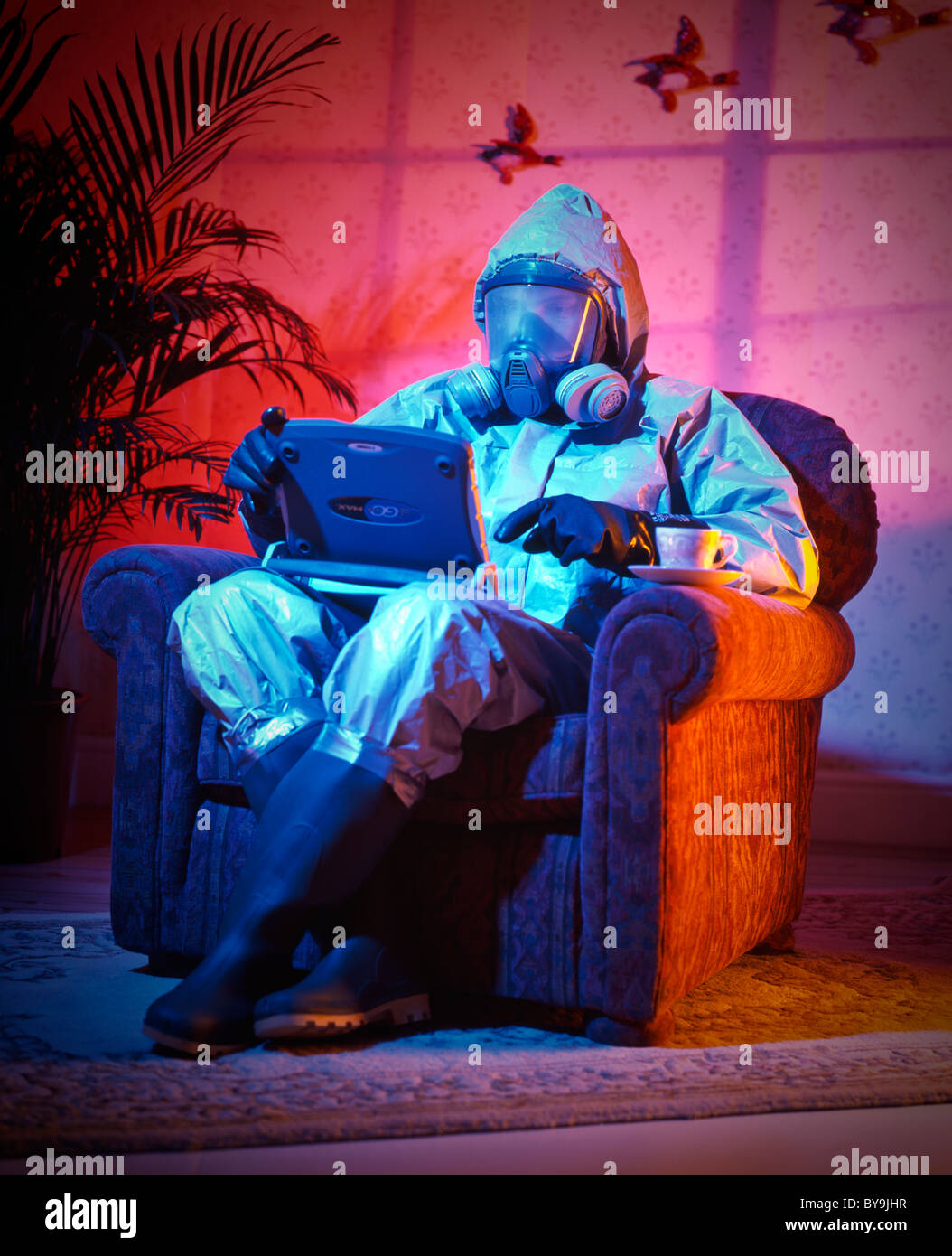 man in sitting in an arm chair in a suburban domestic location with his lap top and a cup of tea wearing a thermo-nuclear suit Stock Photo