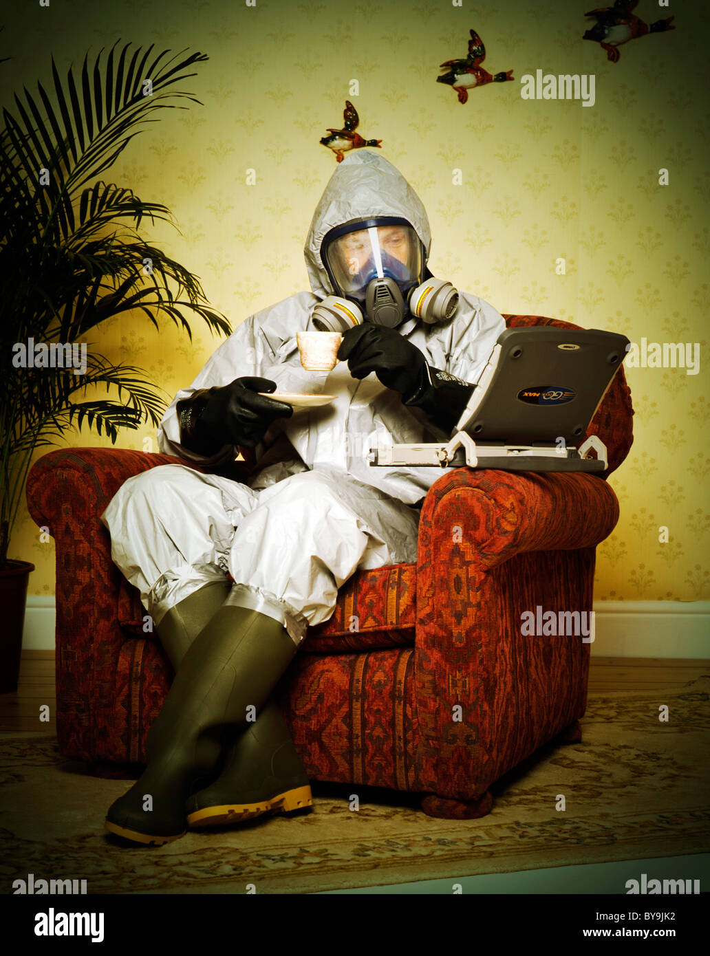 man in sitting in an arm chair in a suburban domestic location with his lap top and a cup of tea wearing a thermo-nuclear suit Stock Photo