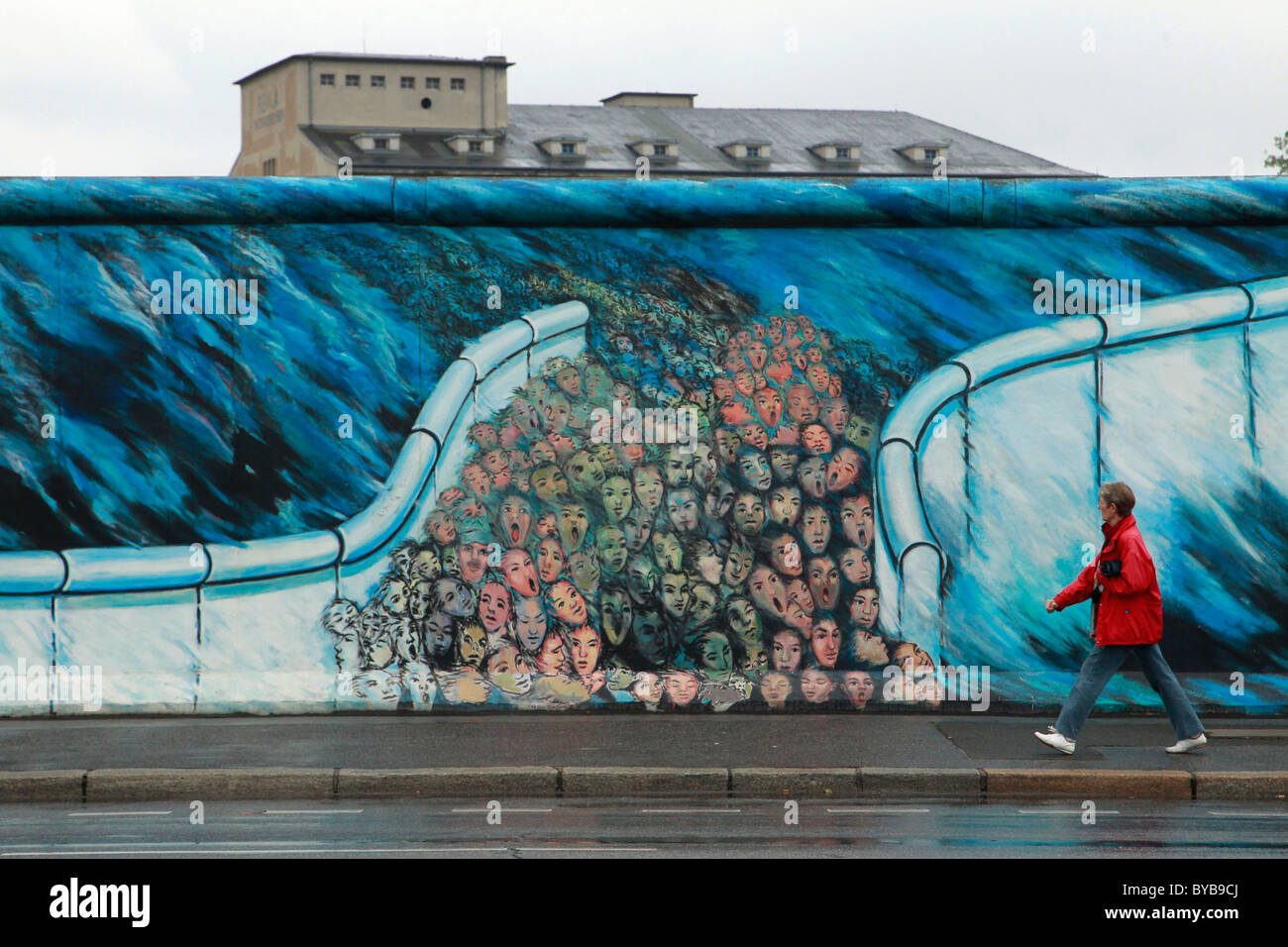 Woman in red jacket passes mural of faces on the Berlin Wall Germany Stock Photo
