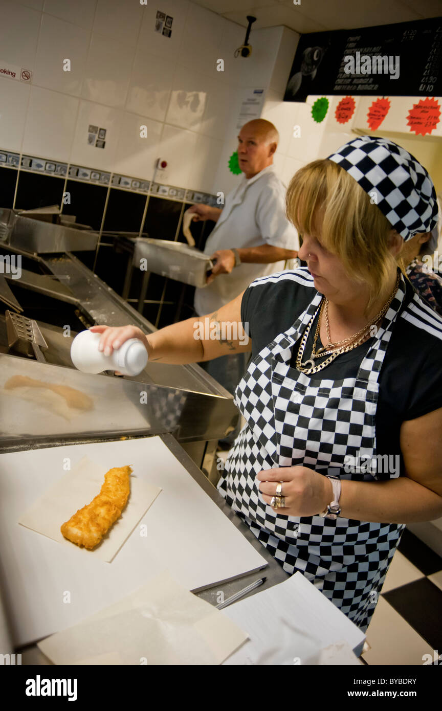 Woman salting a battered fish in a traditional fish and chip shop. A man in the background is frying chips Stock Photo