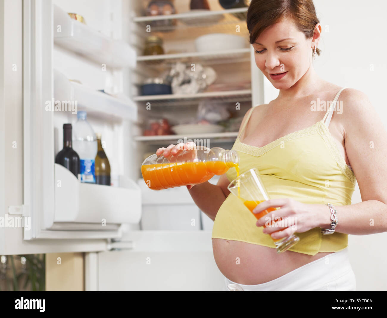 italian 6 months pregnant woman standing near refrigerator and drinking orange juice. Waist up, horizontal shape Stock Photo