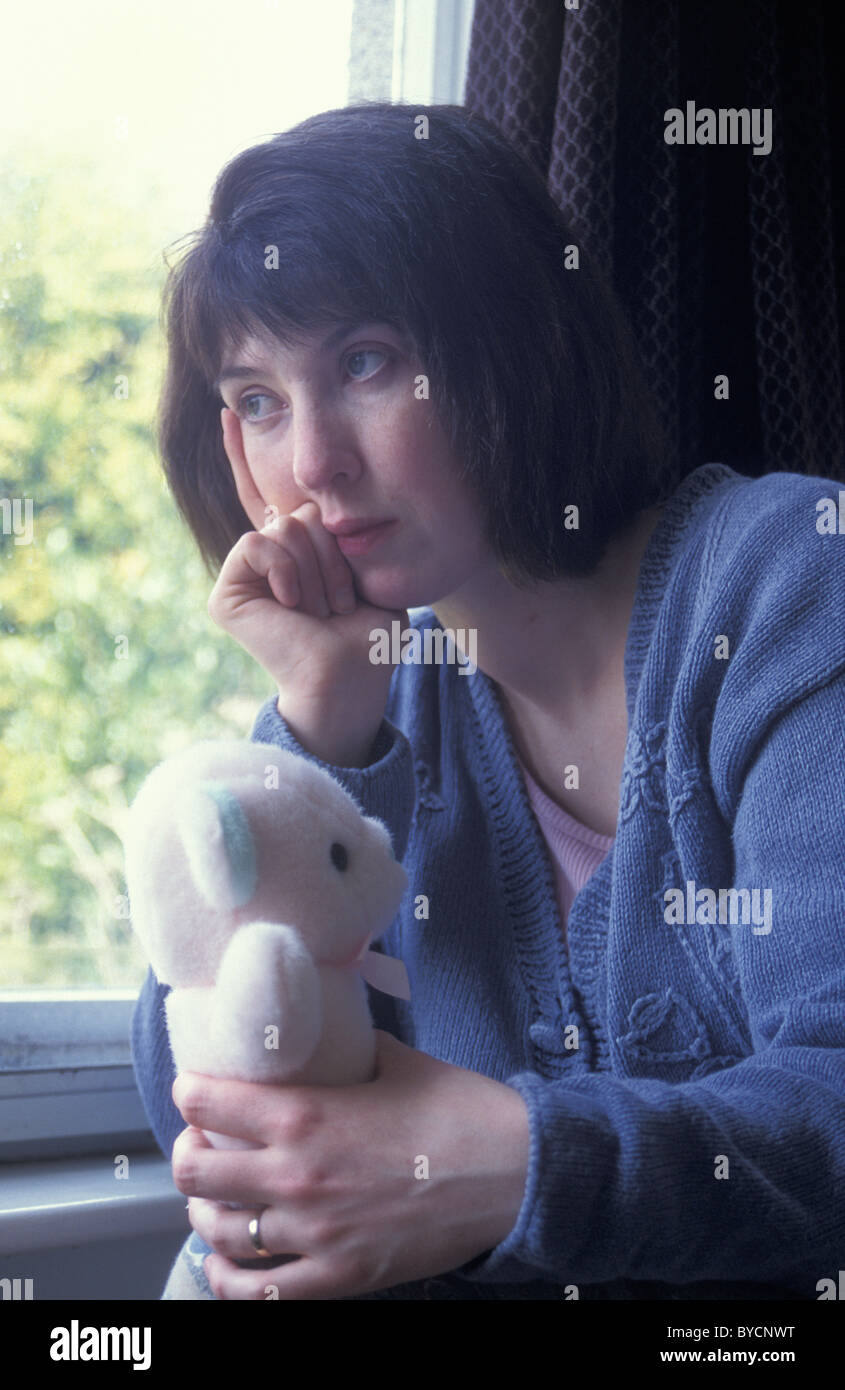 unhappy woman sitting by window holding child's teddy Stock Photo