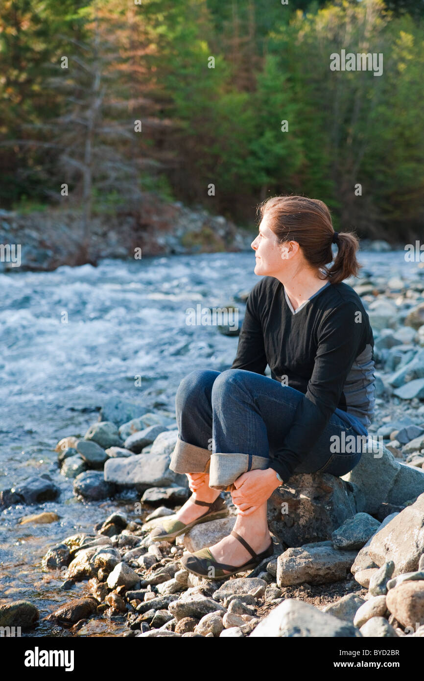 Young woman outside by a rocky river in the sunlight, happy, thoughtful, and content. Stock Photo