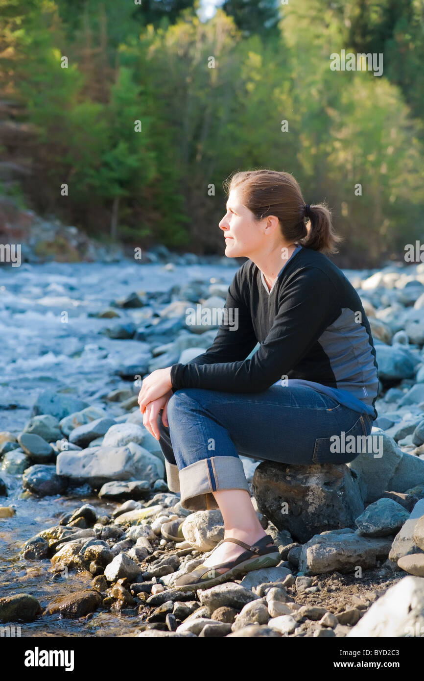 Young woman outside by a rocky river in the sunlight, happy, thoughtful, and content. Stock Photo