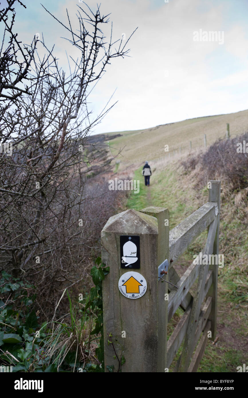 Woman walks along the South West Coastal Path with an open gate in the foreground, Cornwall, England. Stock Photo