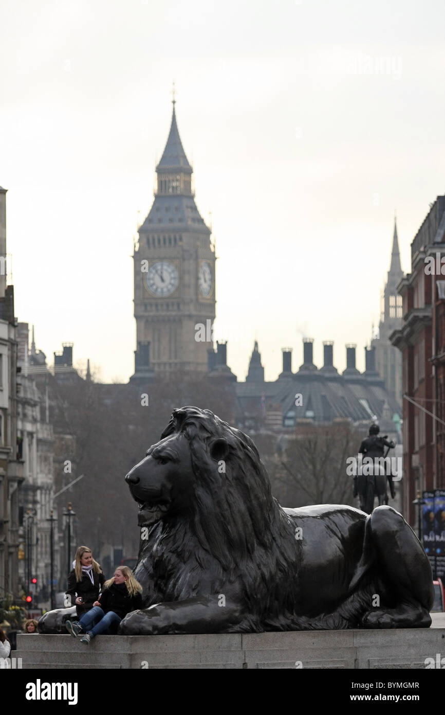 Two girls sitting on the paws of one of the Lions in Trafalgar Square, London, with Big Ben in the background Stock Photo