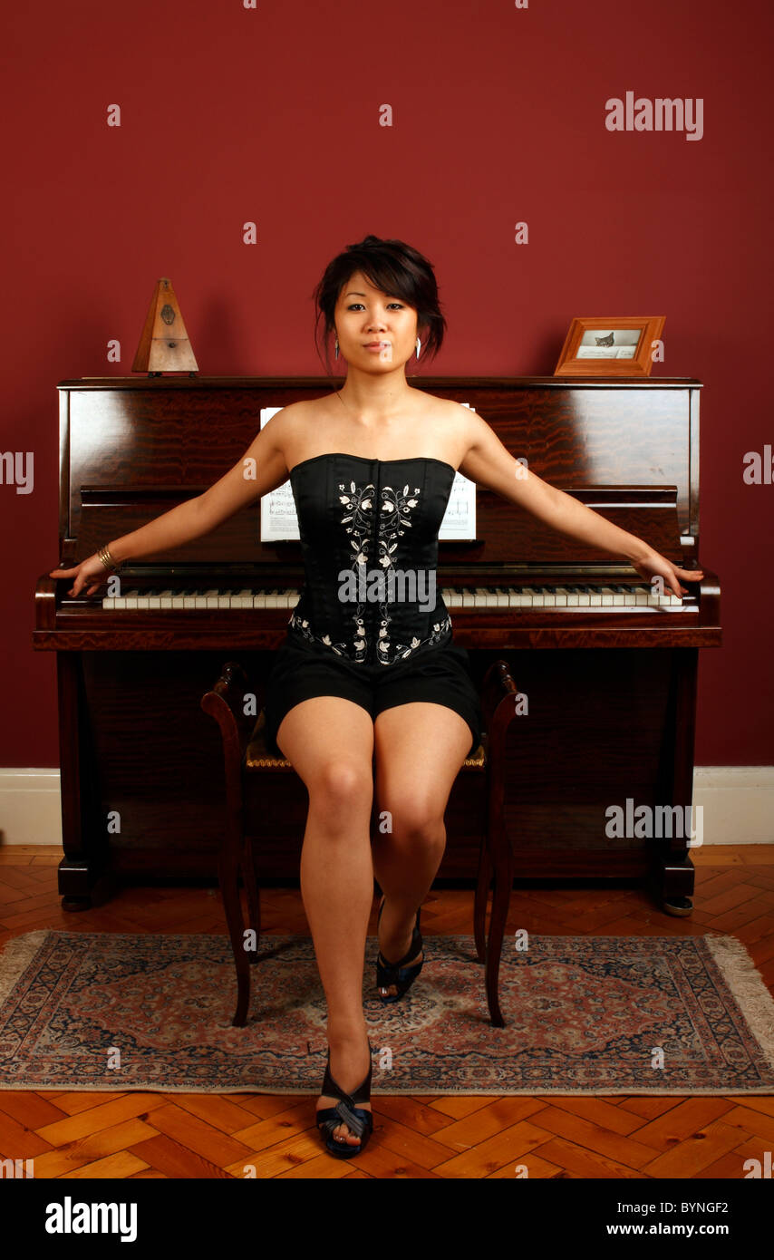 young oriental Chinese woman in a black shorts and an embroidered black basque or corset sitting in front of an upright piano Stock Photo