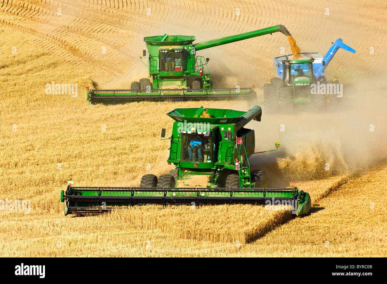 Two John Deere combines in tandem harvest wheat while one unloads into a grain cart “on-the-go” / near Pullman, Washington, USA. Stock Photo
