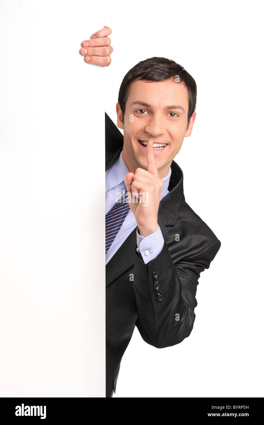 Businessman in a suit gesturing silence with his finger over his mouth, behind white panel Stock Photo