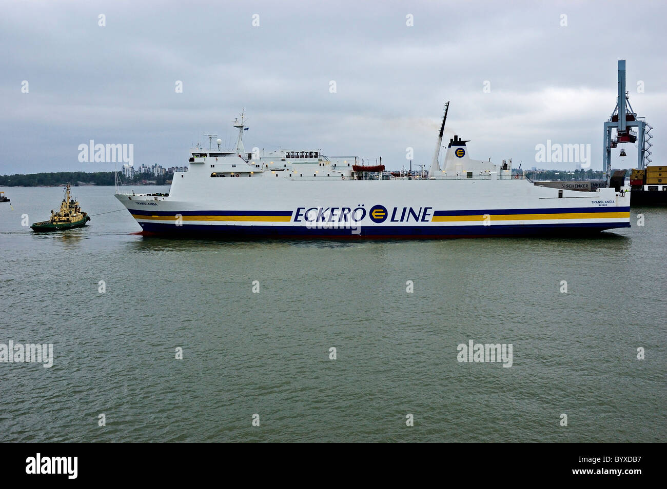 A tug guiding the Eckero ferry Translandia out of harbour towards the green coastline of Helsinki Stock Photo