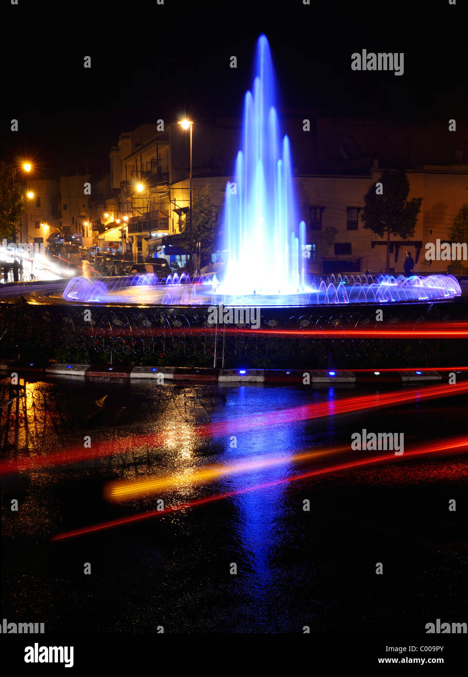 Car taillight streaks and reflections on wet Fes el Bali Medina traffic circle with blue fountain at night Morocco Stock Photo