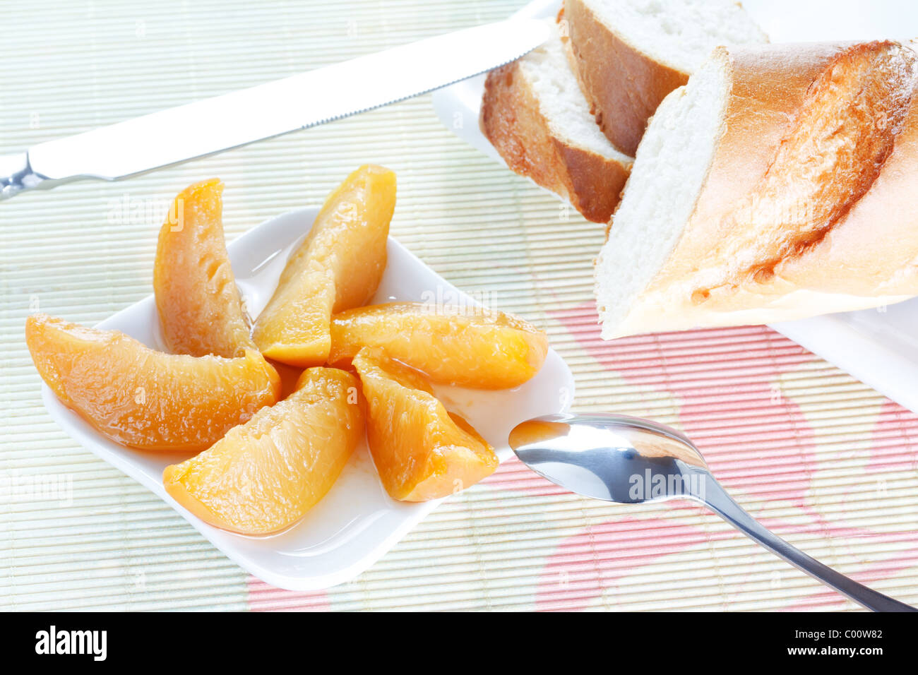 Crisp fresh-baked baguette on white plate, peach dessert, spoon and knife on bamboo placemat Stock Photo