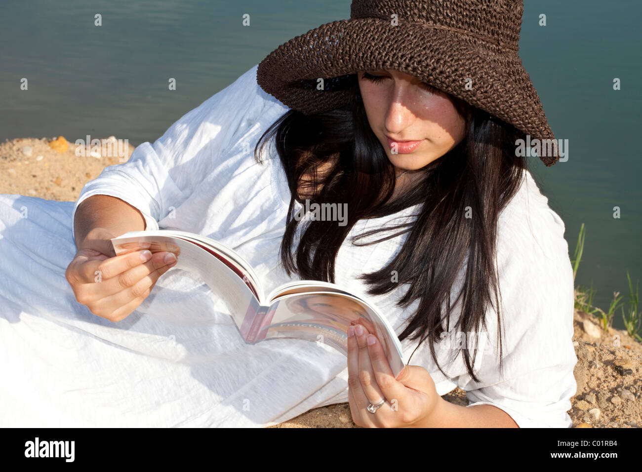 Young woman wearing a straw hat sitting on the beach of a quarry pond ...