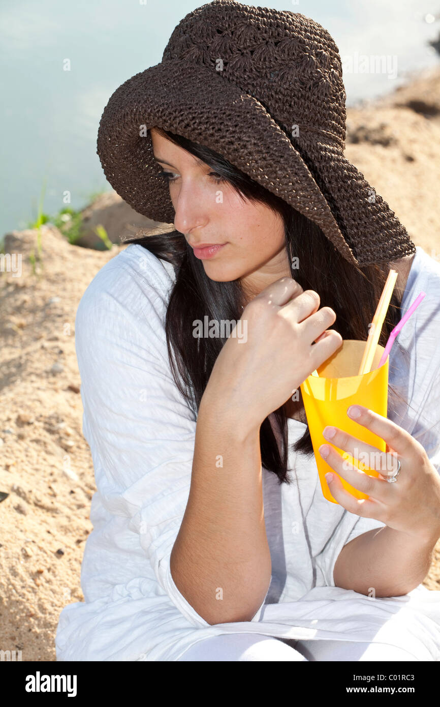 Young woman wearing a straw hat sitting on the beach of a lake and ...