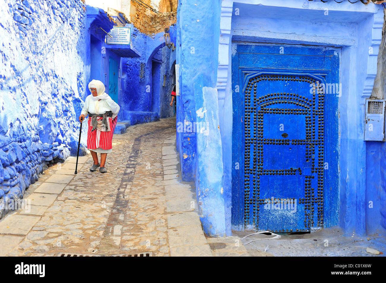 An old woman walking in a narrow alley with blue painted walls and doors in the Medina, old town, Chefchaouen, Rif Mountains Stock Photo